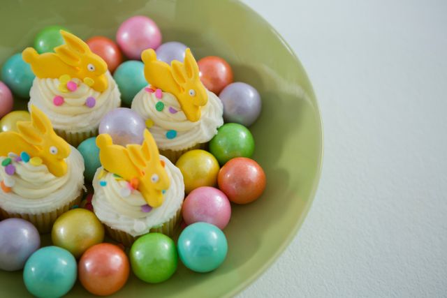 This vibrant image features Easter-themed cupcakes adorned with bunny toppers and surrounded by colorful chocolate eggs in a bowl. Ideal for use in holiday greeting cards, festive party invitations, food blogs, and social media posts celebrating Easter and springtime festivities.