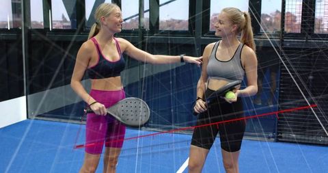 Smiling women playing padel on indoor blue court wearing sports bras and shorts