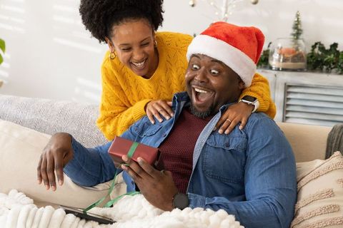 Surprised african american man receiving christmas gift on cozy sofa wearing santa hat
