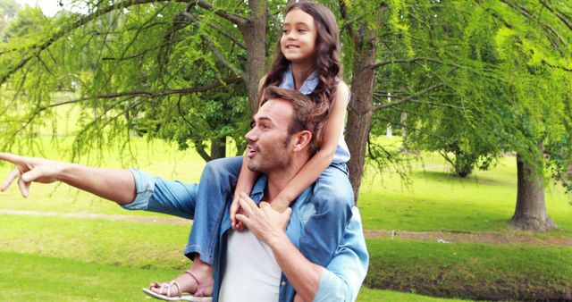 Father carrying daughter on shoulders while pointing at something in the park. Bright, sunny day with lush greenery. Represents family time, bonding, and joy. Ideal for materials about family activities, parenting, outdoor recreation, and happy childhood moments.