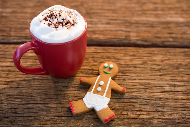 This image shows a red mug filled with coffee topped with whipped cream and sprinkled with coffee powder, placed next to a gingerbread man cookie on a rustic wooden table. Ideal for holiday-themed promotions, Christmas cards, festive blog posts, or social media content celebrating the winter season.