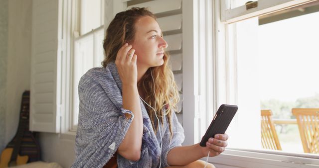 Young woman leisurely sitting by a window, using earphones to listen to music on her smartphone. Ideal for illustrating concepts of relaxation, leisure, modern technology use at home, self-care, and enjoying a sunny day indoors.