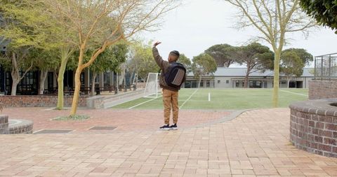 Child Waving in Schoolyard with Backpack and Sports Field