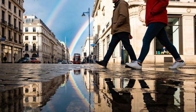 Crossing pedestrians reflecting double rainbow and red bus on wet cobblestone street