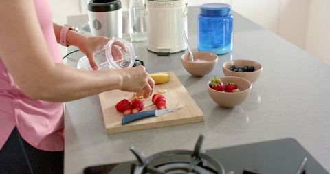 Mature woman preparing fresh fruit smoothie pouring blender cup and slicing strawberries