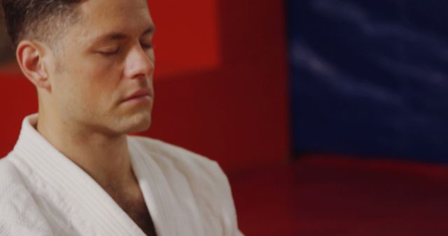 Person in deep meditation moment during martial arts practice, meditating while wearing a white gi in background with contrast of red and blue walls. Useful for topics related to mindfulness, martial arts, meditation, focus, mental health, wellness and personal development.