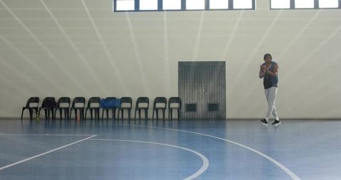Solo man standing on empty indoor basketball court with chairs, duffel and high windows