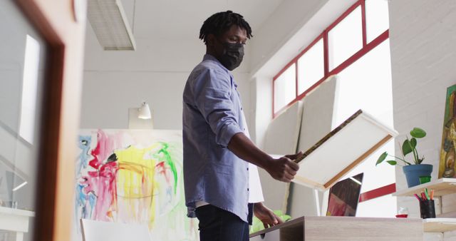 African american male artist wearing face mask holding a painting and using laptop at art studio. art, hobby and creative occupation concept