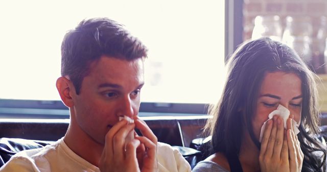 Young couple using tissues, likely suffering from allergy symptoms, sitting together at home. Could be used for articles, advertisements, or blog posts related to allergy relief, health conditions, healthcare products, or home environment improvement.