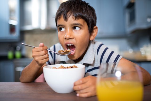 Boy eating breakfast cereal in kitchen, looking away. Ideal for use in articles about healthy eating habits for children, morning routines, or family life. Suitable for educational materials, parenting blogs, and nutrition guides.