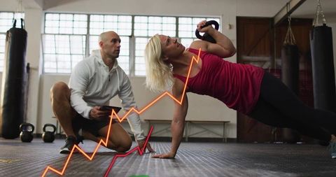 Woman performing kettlebell side plank while trainer coaching with tablet in gym