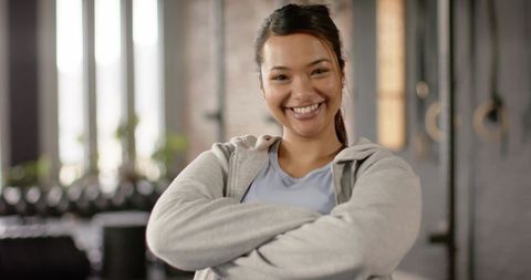 Confident smiling woman crossing arms in gym wearing gray hoodie with kettlebells