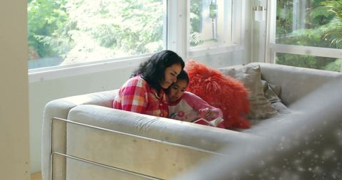 Mother and daughter reading together on sunlit couch with cozy orange accent pillow
