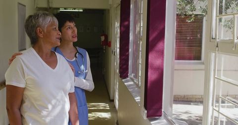 Female Doctor Assisting Senior Woman in Retirement Home Corridor