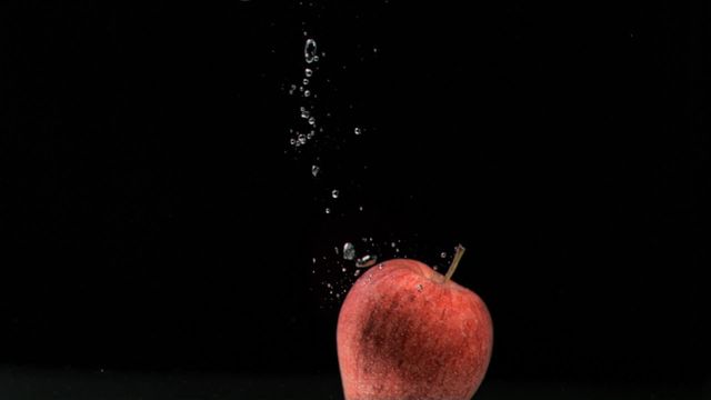 The image captures a red apple in slow motion being immersed in water with droplets creating a splash effect against a black background. Can be used for advertising campaigns focused on freshness, health, organic products, or beverages. Suitable for websites promoting healthy eating or fruit-based recipes.