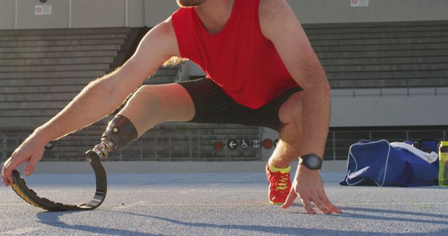 Athlete with prosthetic leg stretching on outdoor track before exercise. Wearing red sleeveless shirt, watch, and running shoes, focusing on flexibility and readiness. Inspirational depiction of overcoming challenges, perfect for use in health, sports, fitness, and motivational campaigns.