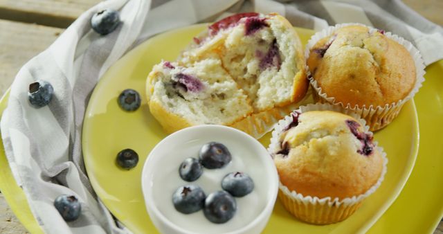 Freshly baked blueberry muffins are displayed on a yellow plate, accompanied by a small dish of blueberries. The vibrant colors and close-up view invite a sense of home-baked warmth and the simple pleasure of a sweet treat.