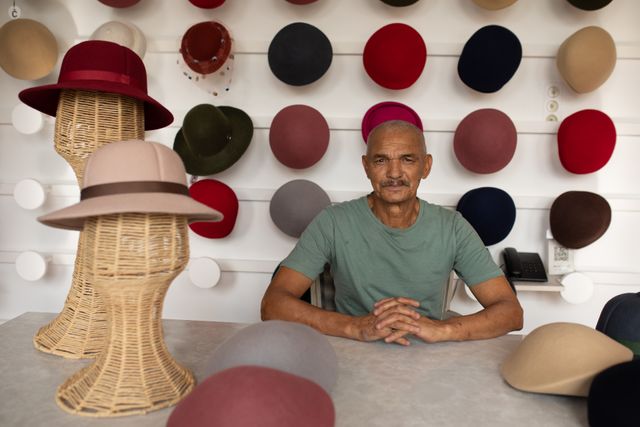 Portrait of a happy senior biracial man sitting at a table and surrounded by hats on display in the showroom at a hat factory.