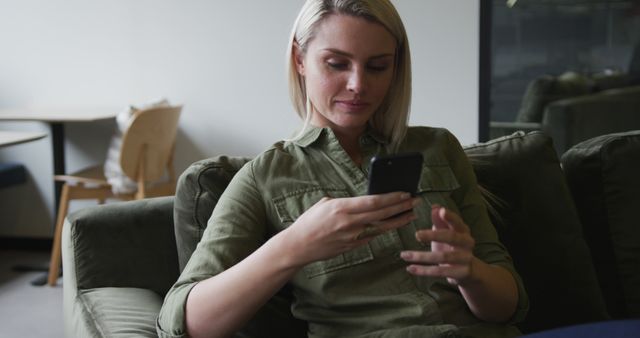 Young woman with blonde hair using a smartphone while relaxing on a comfortable couch in a contemporary living space. Perfect for advertising casual lifestyle, home decor, technology products, or social media content.