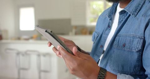 African american man using tablet in bright modern kitchen with denim shirt, wristwatch