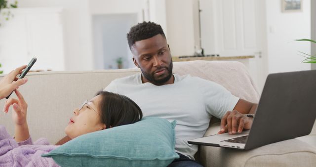 Mixed race couple relaxing on sofa; one on laptop, other on smartphone. Great for themes of home life, relaxation, technology use in modern lifestyle, and togetherness in domestic environment.