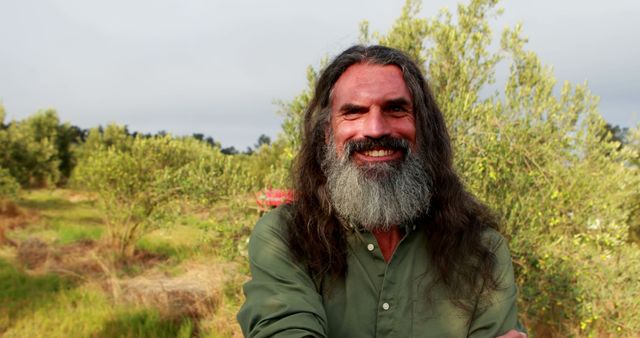 Smiling man with long hair and beard stands in olive grove, perfect for depicting lifestyle, nature, relaxation themes. Ideal for use in advertisement, editorial content, website banners, and health and wellness promotions.