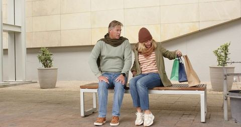 Mature couple relaxing on bench holding shopping bags in urban plaza