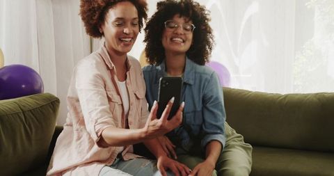 Smiling friends taking selfie on green sofa with smartphone, balloons and cozy home vibe