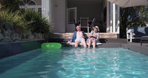 Mature couple relaxing at backyard poolside, dipping feet in sunlit water, summer lifestyle