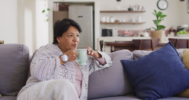 Mature woman sitting on a comfortable sofa, enjoying a beverage in a cozy and casual living room. Scene conveys relaxation and comfort, perfect for use in lifestyle blogs, wellness articles, and advertisements promoting home comfort products.