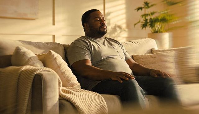 Middle-aged man relaxing on cozy beige sofa in warm sunlight closing eyes for mindfulness