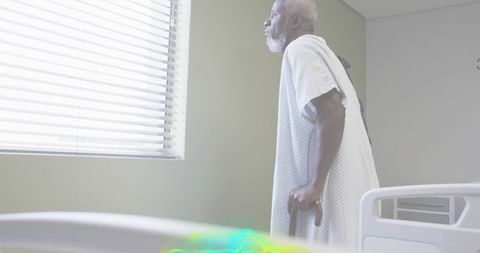 Sunlit senior standing in hospital gown leaning on cane beside bed for recovery and reflection