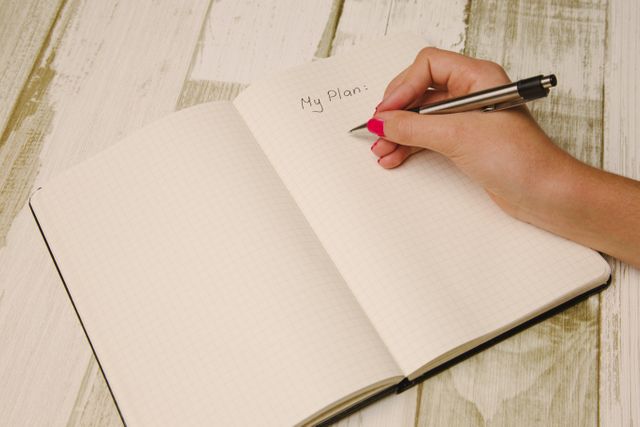 Close-up shot of a female hand with red nail polish writing 'My plan' in a journal on a white wooden table. Ideal for use in articles or blog posts about planning, productivity, organization, or bullet journaling. Useful for websites promoting stationery, notebooks, and productivity tools.