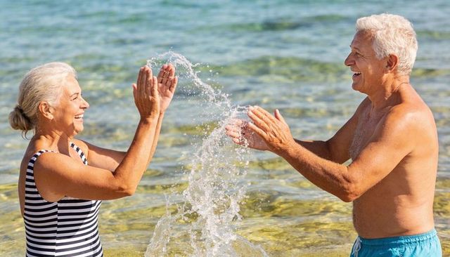 Senior couple splashing water and laughing at sunny seaside, active retirement lifestyle