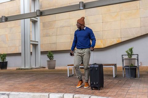 Urban traveler standing with rolling suitcase and headphones outside transit hub