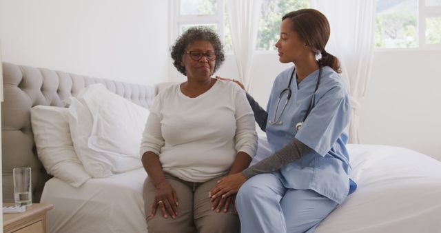 Senior african american woman and female doctor sitting on bed, talking. Medicine, healthcare and lifestyle.