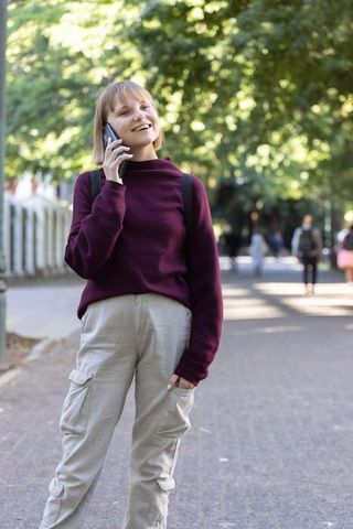 Young woman talking on smartphone while walking under sunlit tree canopy on city pathway