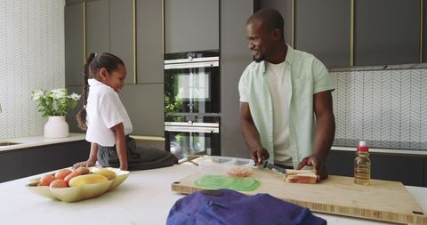 Diverse father preparing school lunch with smiling daughter on modern kitchen island