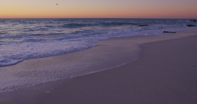 Gentle waves lap at sandy beach as the sun sets, casting a warm glow over the horizon. Perfect for projects about relaxation, travel destinations, nature appreciation, backgrounds for websites focused on wellness, and promotional material for seaside resorts.