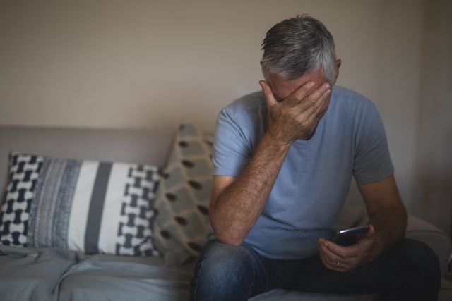 This image depicts an older man experiencing stress or anxiety while holding a mobile phone and sitting on a sofa at home. It can be used in articles or advertisements related to mental health, senior care, technology use among older adults, or emotional well-being. The setting suggests a personal and intimate moment, making it suitable for campaigns focused on mental health awareness or support services for seniors.