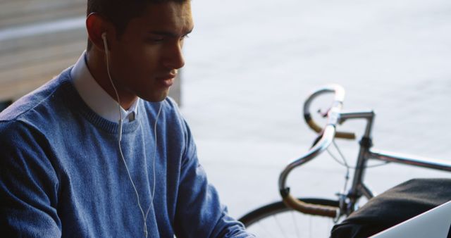 Young man wearing casual outfit is working on his laptop outside, near a bicycle. This can be used for promoting work-from-home culture, urban living, outdoor productivity, freelance lifestyle, or connectivity.