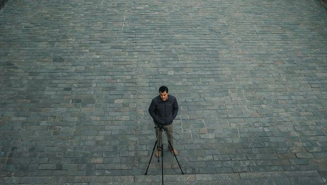 Adjusting photographer checking tripod and camera on empty urban stone plaza from above