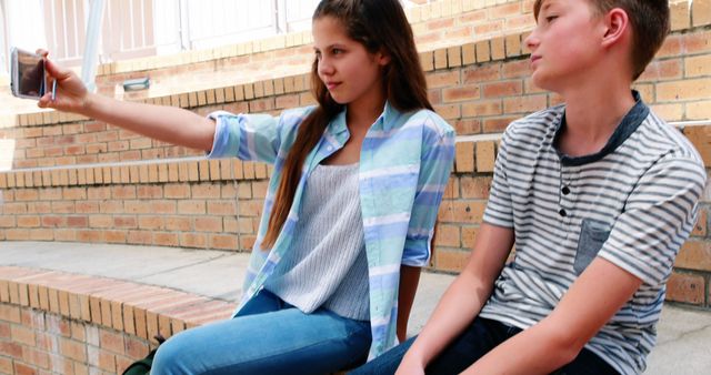 A teenage girl and boy are sitting together outdoors as the girl takes a selfie with her phone, with copy space. Capturing moments of friendship and technology use among teens, the image reflects contemporary youth culture and social media trends.