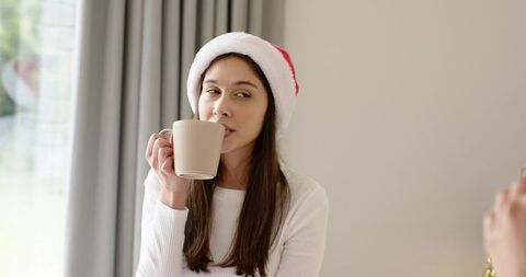Young woman wearing santa hat sipping warm mug by window in cozy morning light