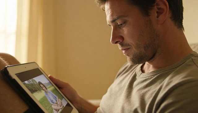 Man viewing woman on tablet while relaxing on couch by soft sunlit curtain, casual tech