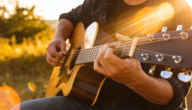 Playing acoustic guitar in golden-hour meadow with sunflare and warm bokeh light, close-up