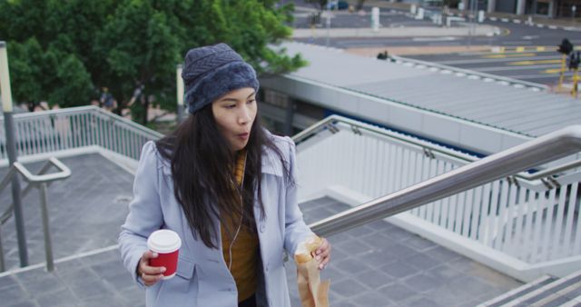 Asian woman walking holding takeaway coffee and eating sandwich. digital nomad on the go, out and about in the city.