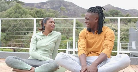 Diverse couple practicing yoga and chatting on outdoor deck with mountain and greenery backdrop
