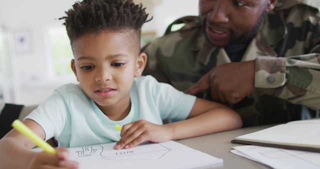 A military father in uniform helps his young son with homework at home. This image showcases family bonding, support, and the importance of education. It highlights the strong relationship between a parent and child and can be used in articles or advertisements related to military families, early childhood education, parental support, or family life.