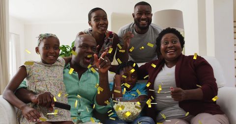 a joyful multigenerational family sitting together on the couch celebrating a sports game with gold confetti in the air. this captures the spirit of togetherness and excitement during family time. suitable for use in promotions about family products, home entertainment, or sports entertainment content.